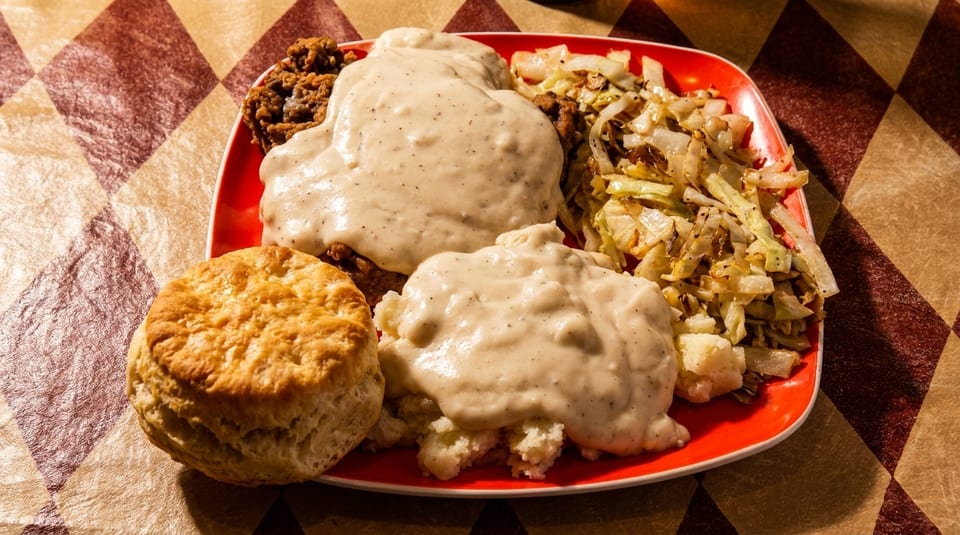 A red plate holds chicken fried steak with gravy, mashed potatoes, fried cabbage, and a biscuit.