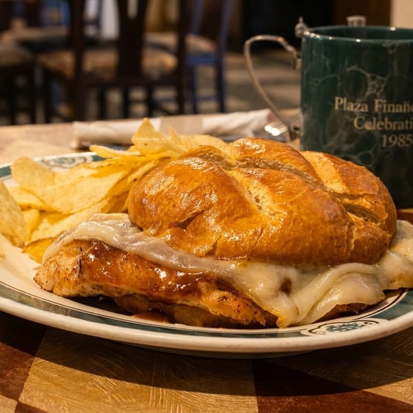 A chicken sandwich with melted cheese and chips on a plate, next to a green mug.