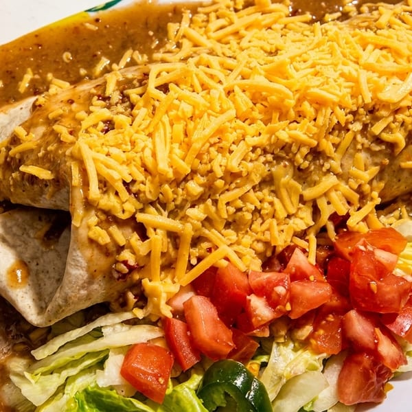 A large burrito covered in green chili sauce and shredded cheese, with a side of lettuce and diced tomatoes.