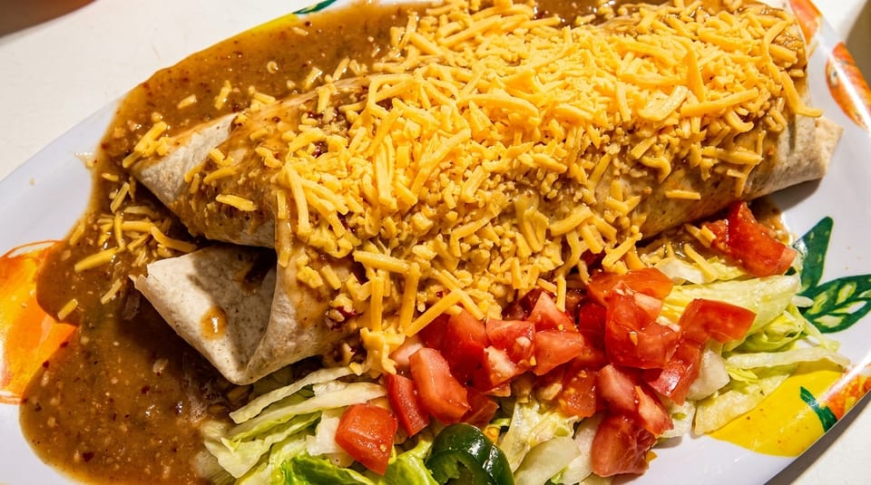 A large burrito covered in green chili sauce and shredded cheese, with a side of lettuce and diced tomatoes.